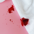 Gotas de sangue vermelho escuro espalhadas sobre um tecido branco, com um fundo rosa.