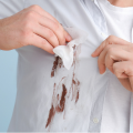 Mãos limpando uma mancha de terra marrom em uma camisa branca com um lenço de papel.